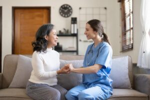 A nurse sits on a couch next to an elderly woman. They are looking at each other and holding hands.